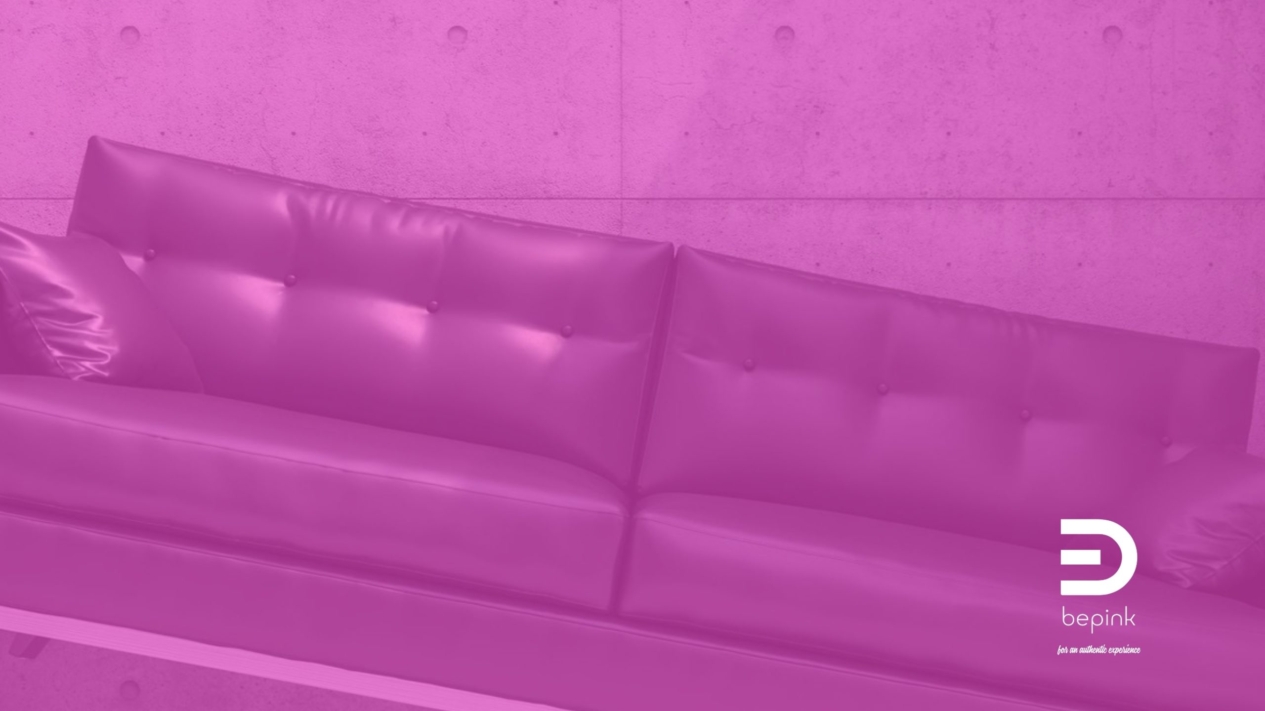 Anatomia dell’immaginazione — Spazi Un divano vuoto con rivestimento viola, davanti a una parete dalla texture materica, in un ambiente dominato da tonalità rosa e magenta.